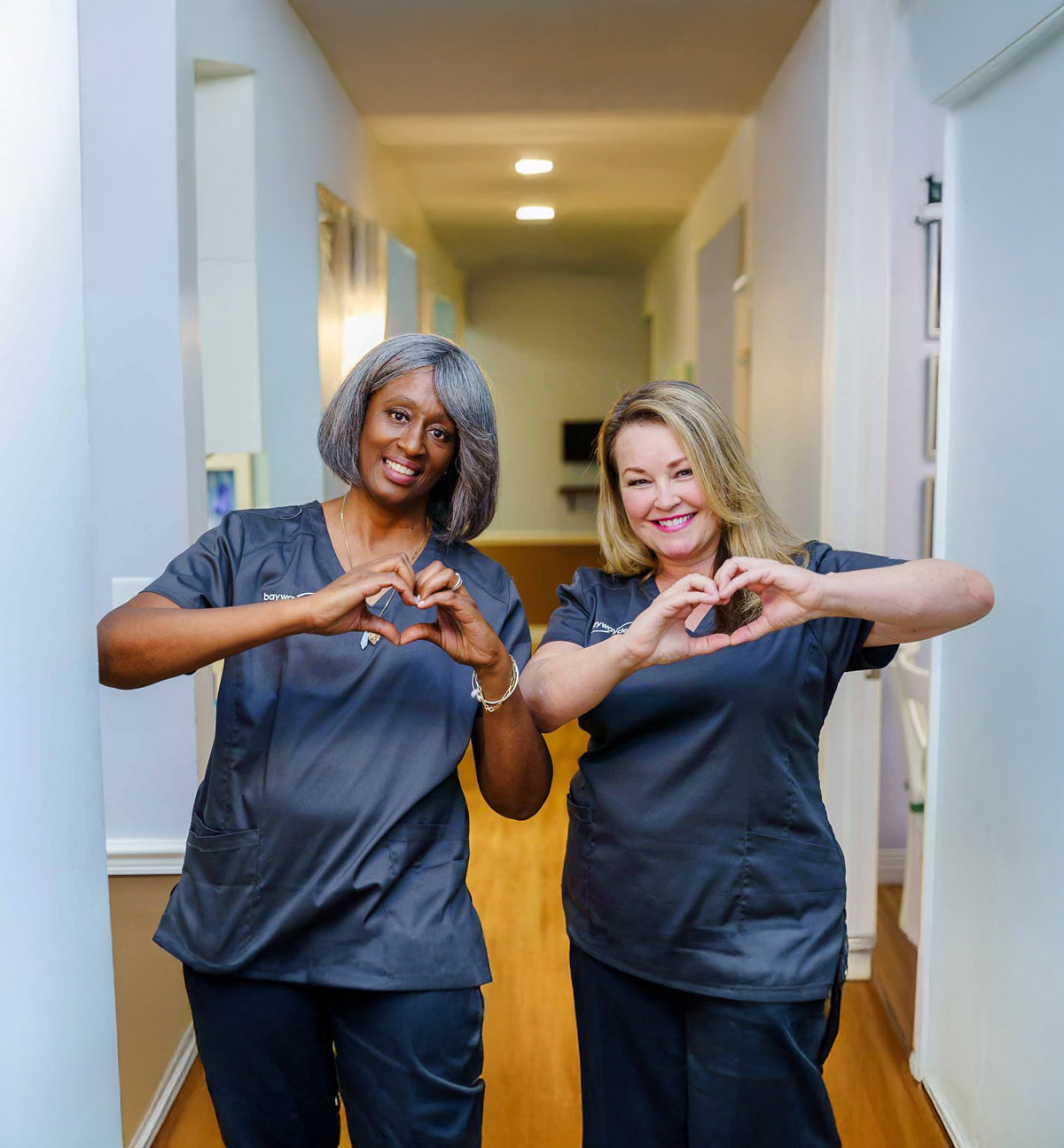 Two women in healthcare uniforms forming heart shapes with their hands, smiling in a brightly lit hallway.